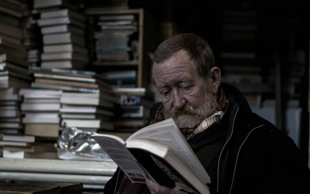 Älterer Mann mit einem Buch in der Hand, im Hintergrund Bücherstapel